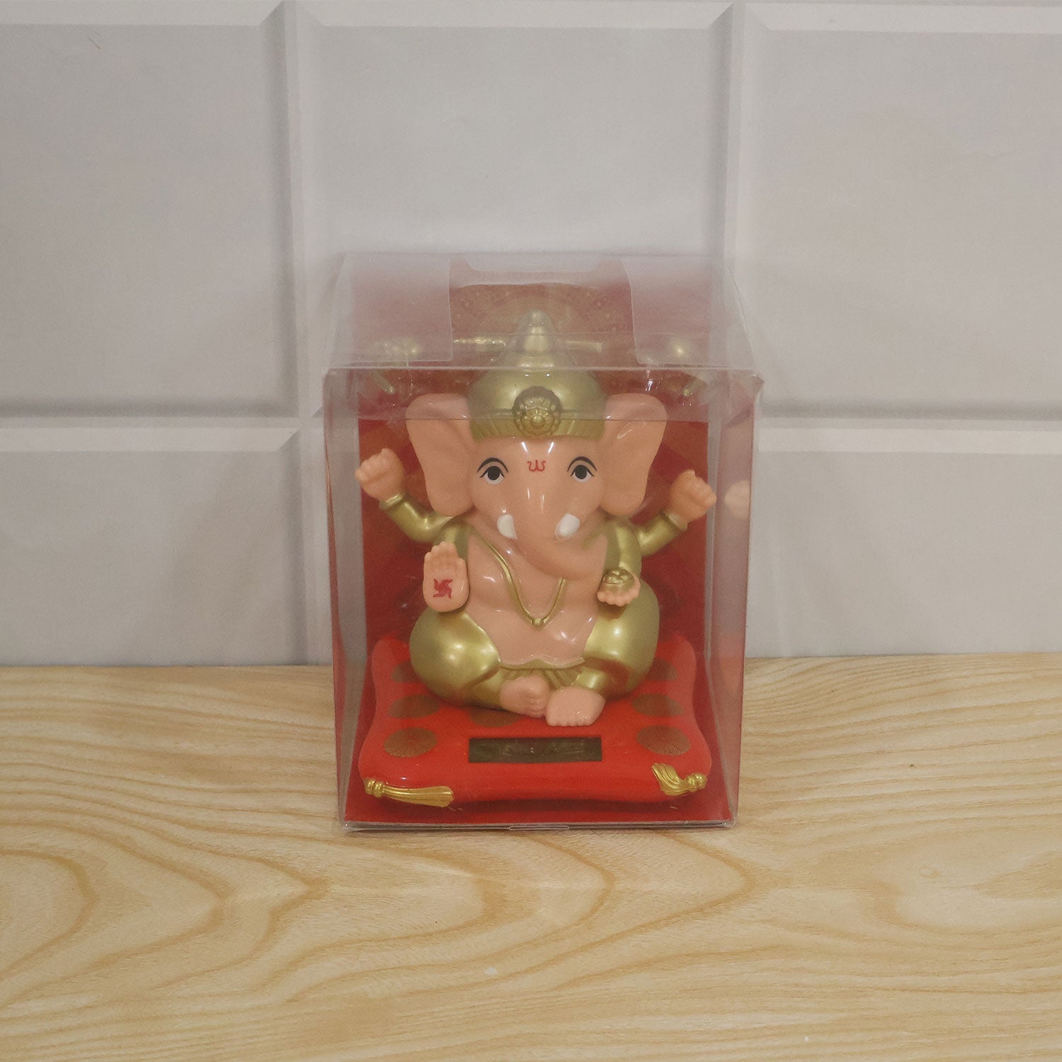 A peach and gold Ganesha idol on a red cushion with a solar panel, packaged in a clear plastic box.