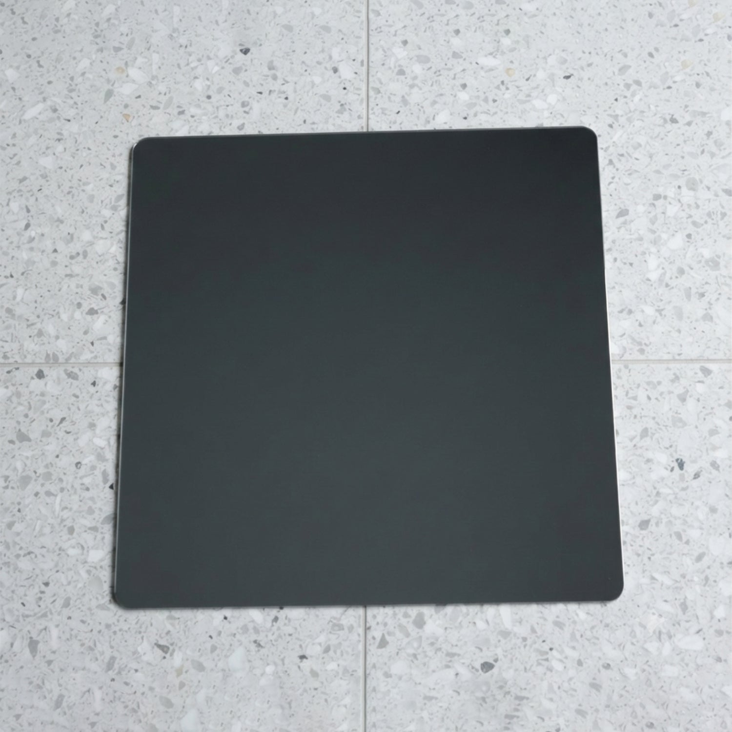 A square mirror with rounded corners lies flat on a speckled gray and white tiled floor.