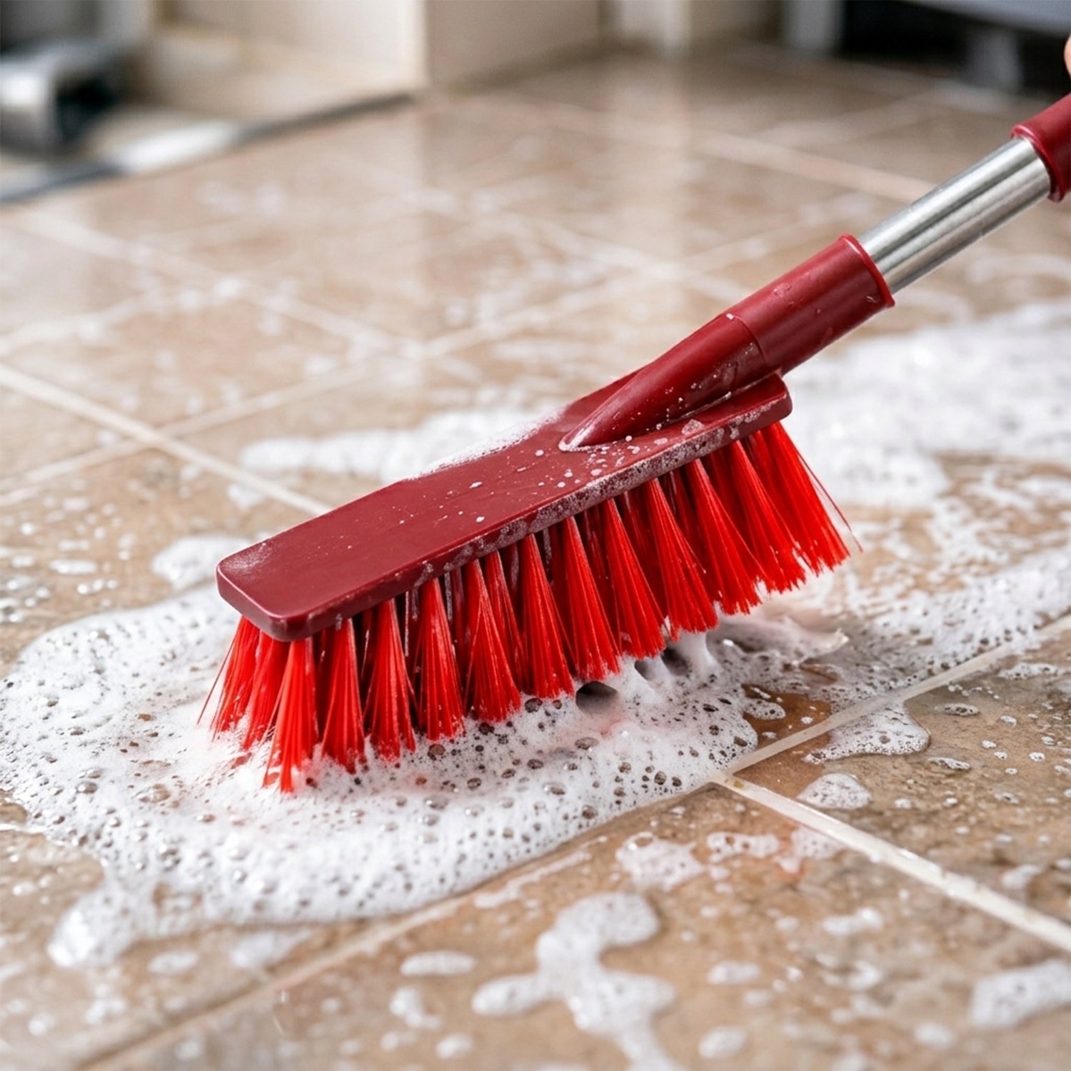 A red and silver brush with red bristles is scrubbing a tiled floor covered in white soap suds.