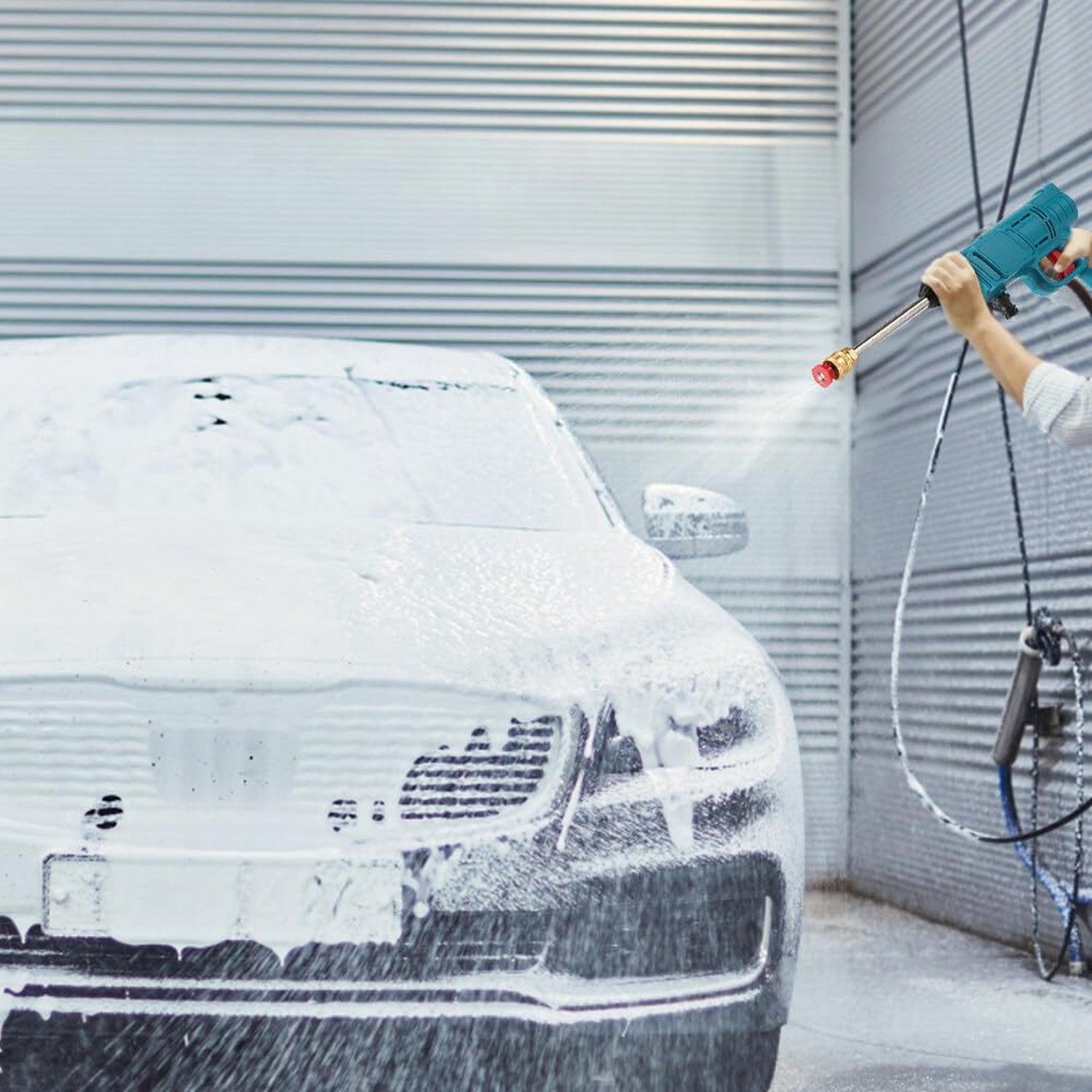 A person's hands hold a teal pressure washer, spraying water onto a white foamy car in front of a gray garage door.