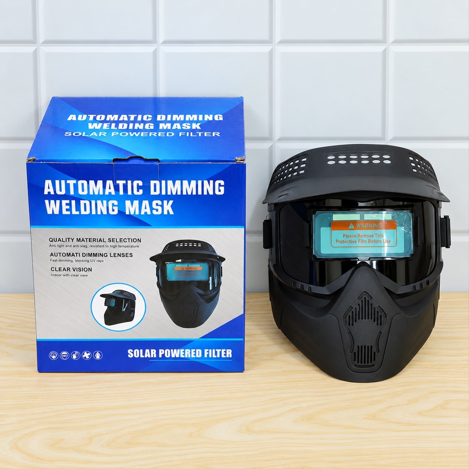 The black welding helmet is displayed next to its blue product box, which features text and images of the mask.