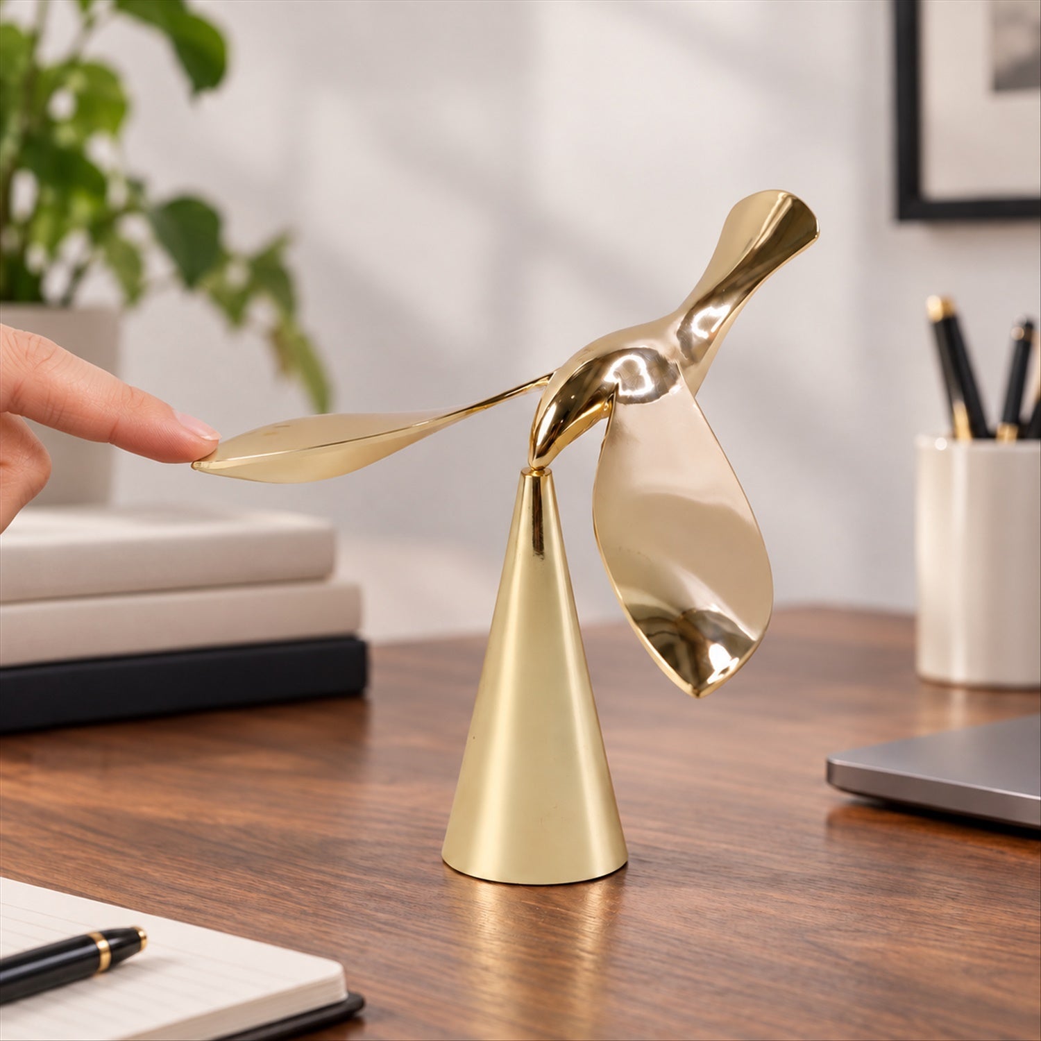 A finger gently touches the wing of a gold balancing bird sculpture on a wooden desk with a laptop nearby.