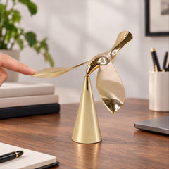 A finger gently touches the wing of a gold balancing bird sculpture on a wooden desk with a laptop nearby.