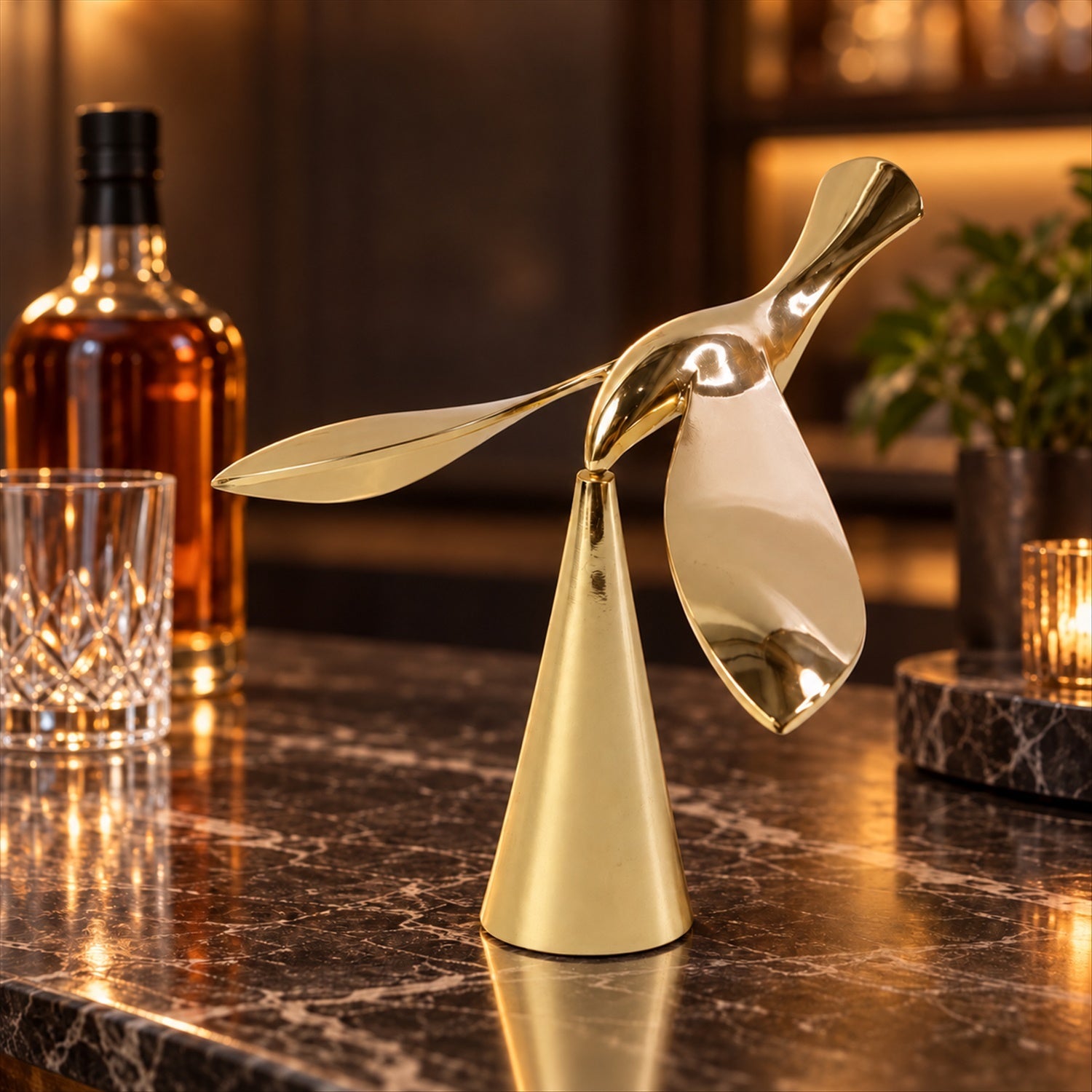 A gold balancing bird sculpture stands on a dark marble bar counter with a whiskey bottle and glass in the background.