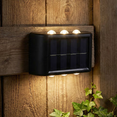 A black solar-powered LED wall light illuminates a wooden fence and green ivy at night.