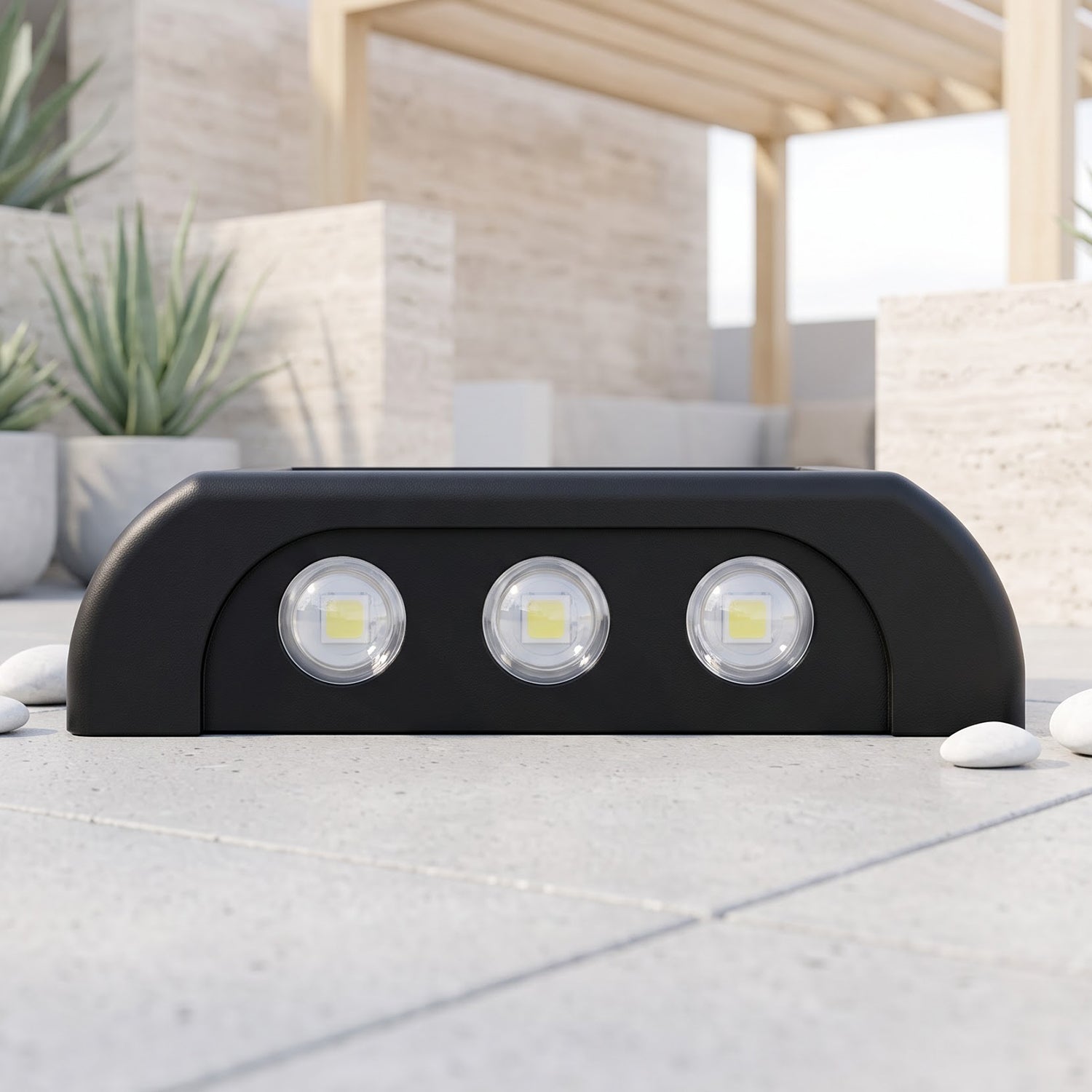 A black solar-powered LED wall light with three bright white LEDs on a light grey tiled patio with white pebbles and plants.