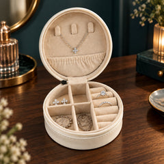 An open cream-colored round jewellery box filled with various silver pieces on a wooden dresser.