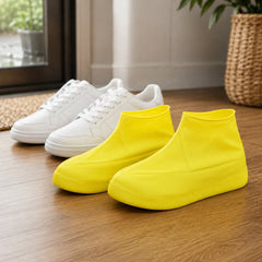 A pair of white sneakers and a pair of bright yellow silicone shoe covers are displayed on a light wooden floor.