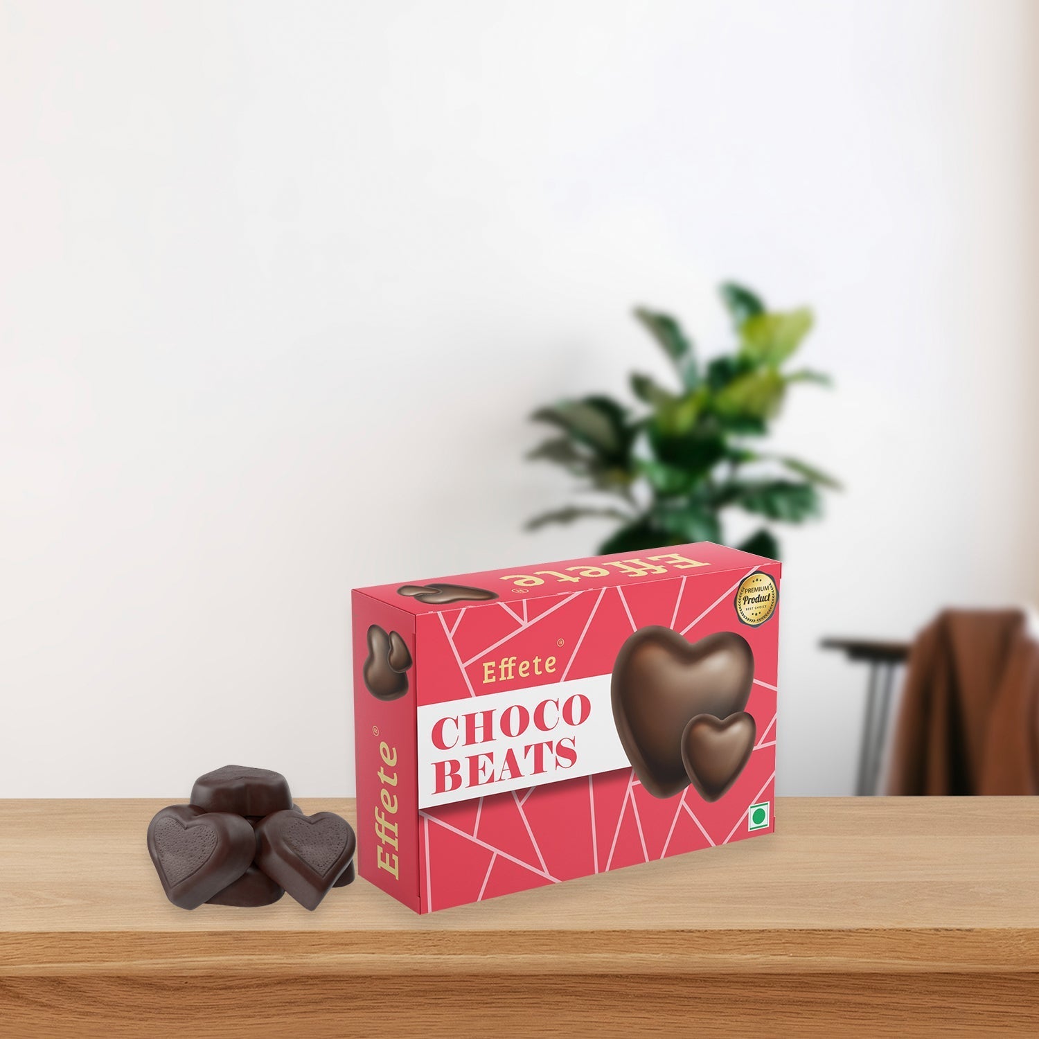 Pink box of heart-shaped chocolates with several loose candies on a wooden table, plant in background.