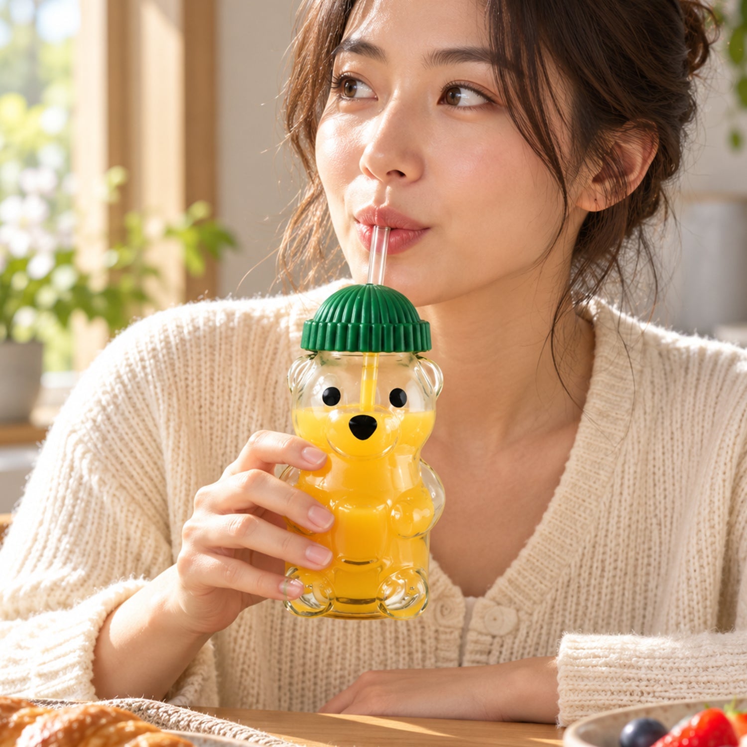 A woman sips orange juice from a bear-shaped glass with a green lid and straw at a sunny table.
