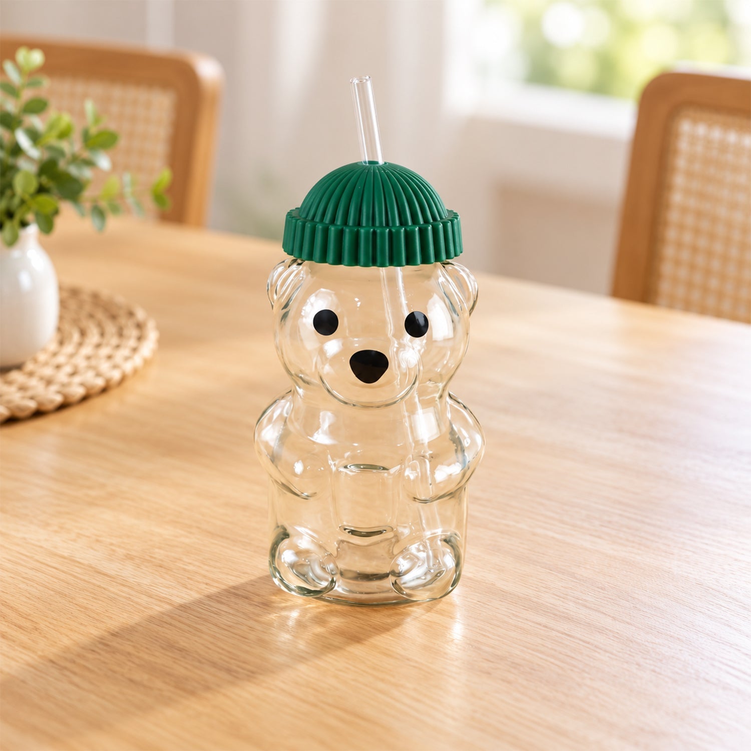 An empty bear-shaped glass sipper with a green ribbed lid and a clear straw sits on a wooden table.