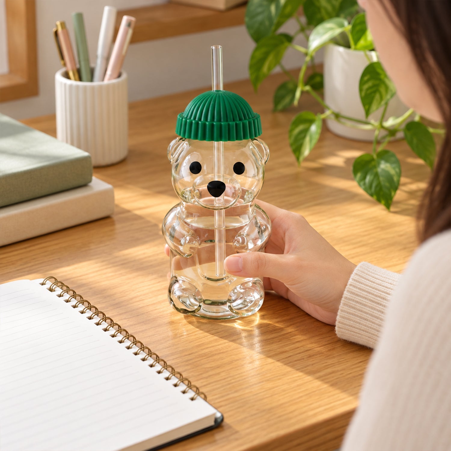 A person's hand holds a bear-shaped glass filled with water on a wooden desk with a notebook and plant.