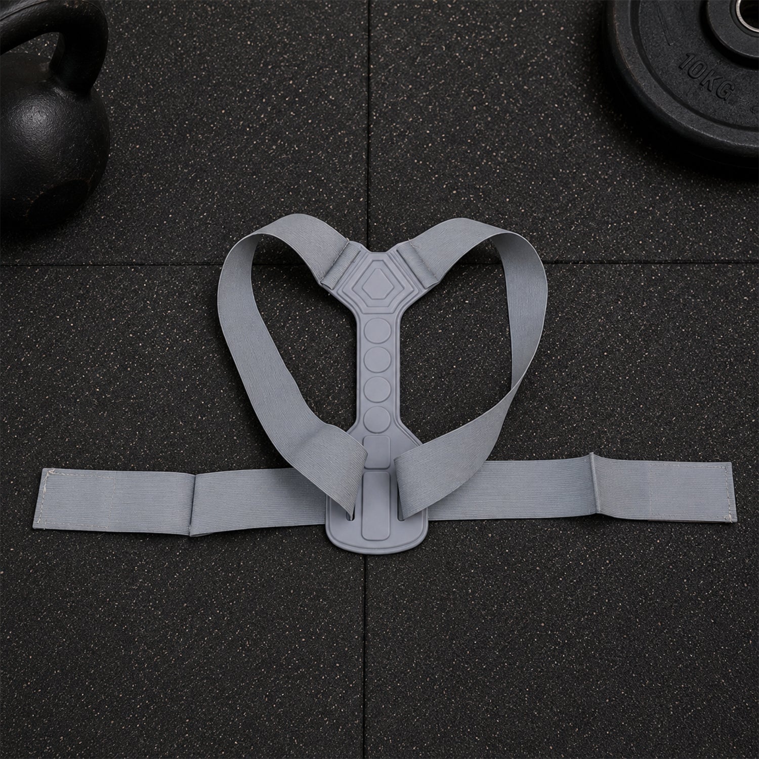 A grey posture corrector belt with a central spine support and shoulder straps laid flat on a dark textured surface.