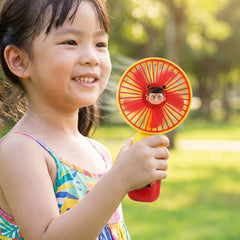 A young girl smiling and holding a red and yellow mini fan with a cartoon character face in the center. â€“ view 3
