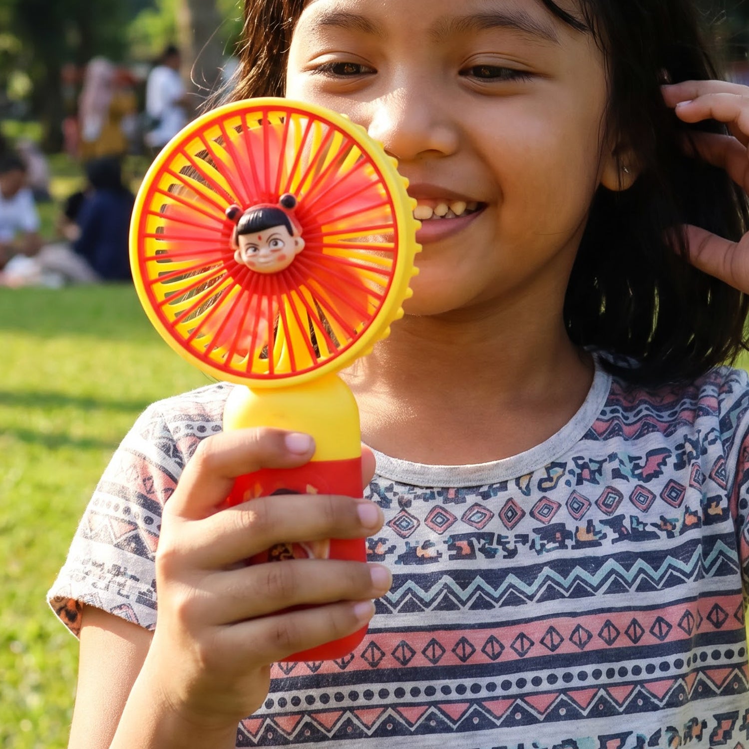 A young girl smiling and holding a red and yellow mini fan with a cartoon character face in the center. â€“ view 7