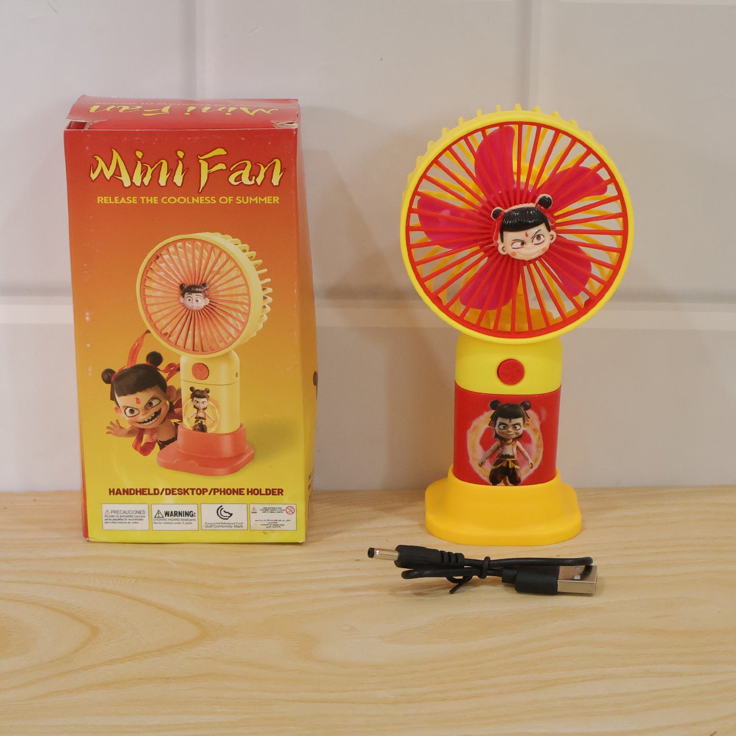 A red and yellow mini fan and its packaging box, with a USB charging cable on a wooden surface.