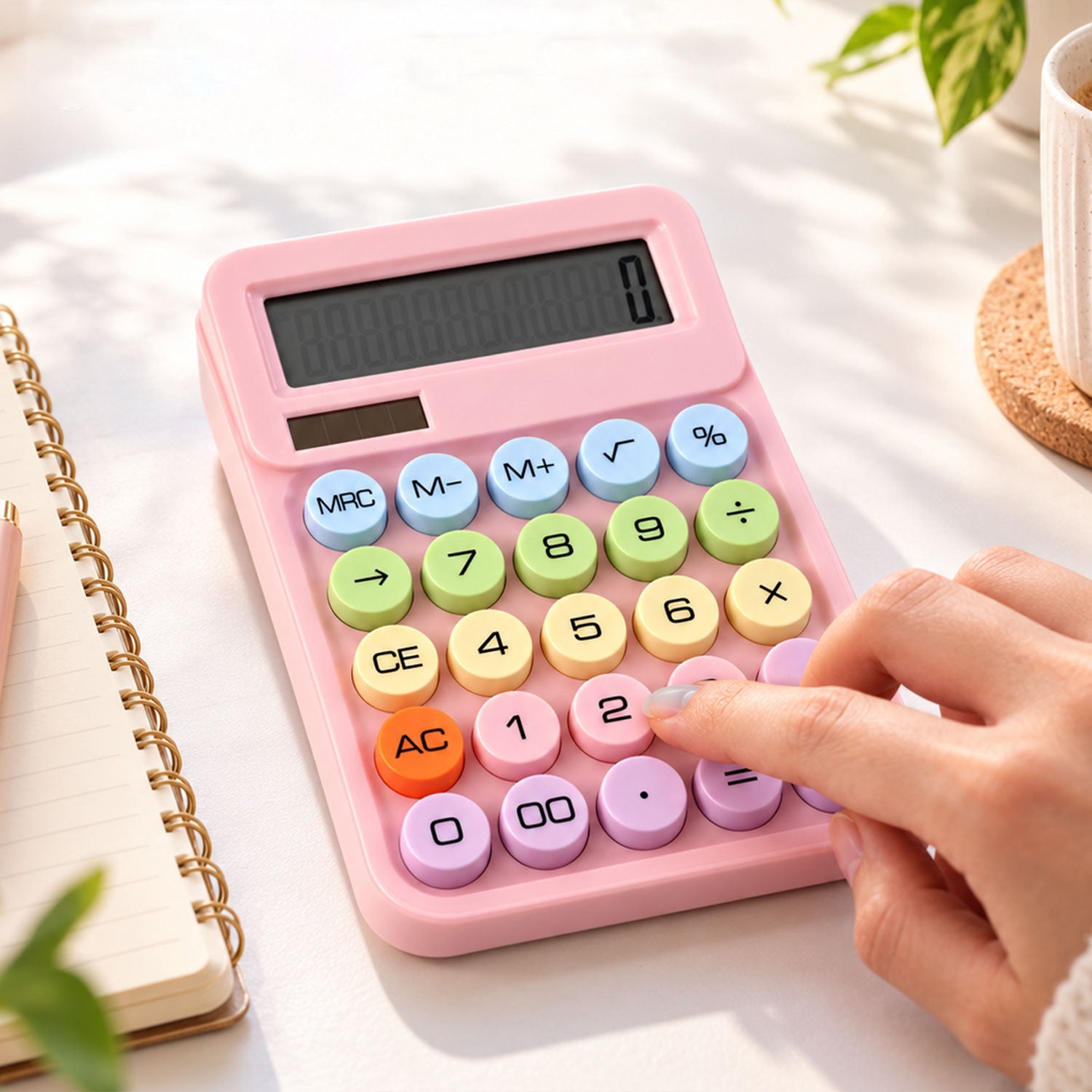 A person's hand presses a pastel pink calculator with multicolored round buttons on a white desk with a notebook and plant.