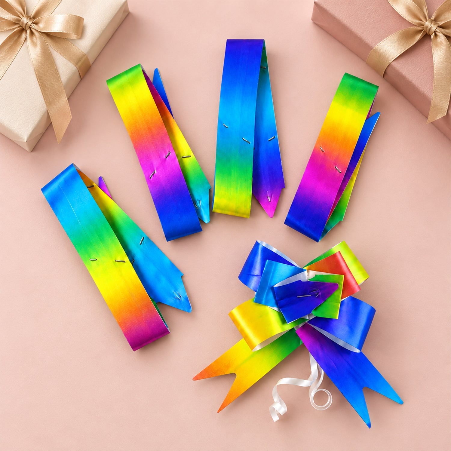 Four flat rainbow ribbons and one assembled rainbow pull bow are displayed on a light pink surface with gift boxes.