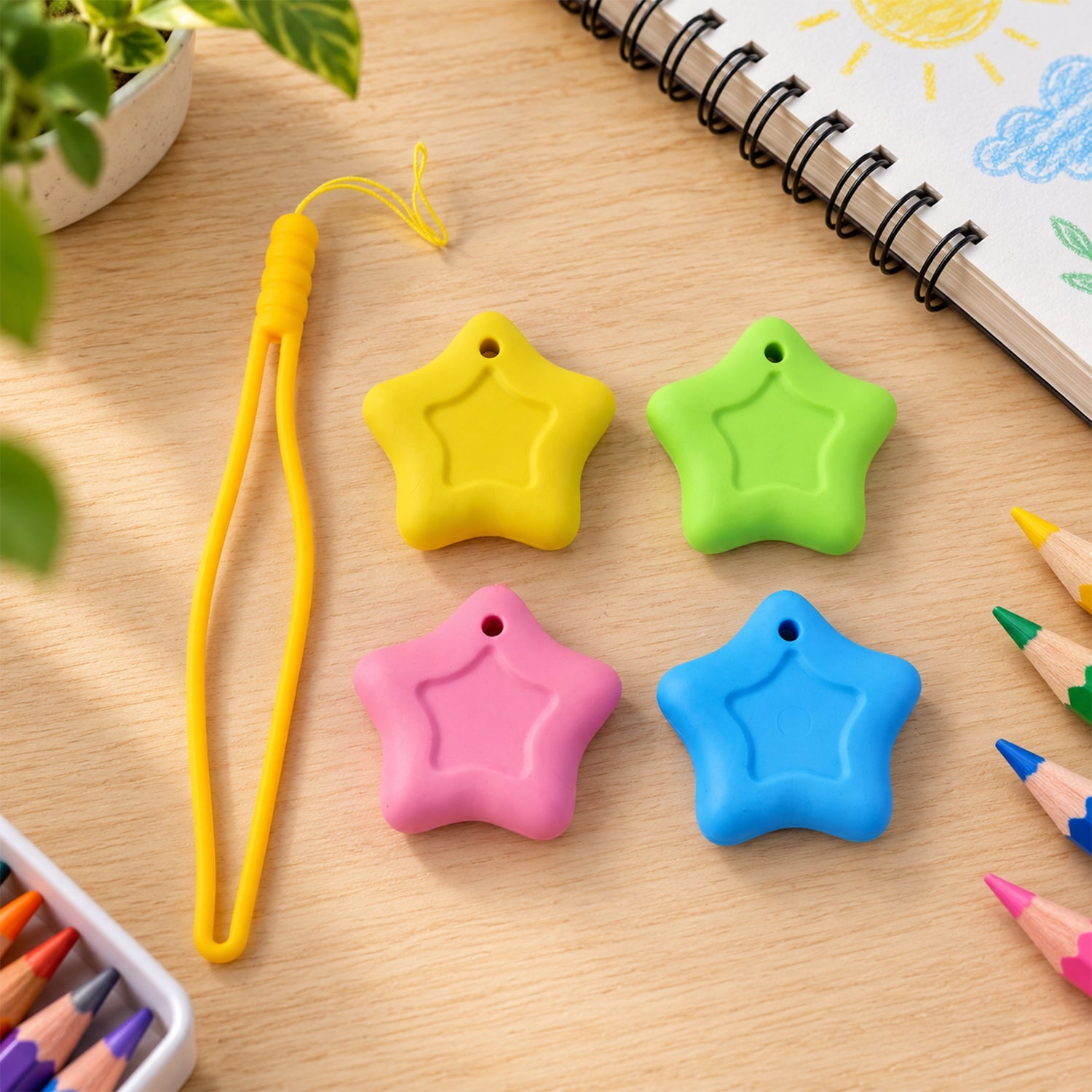 Four star-shaped erasers in yellow, green, pink, and blue sit on a wooden desk next to a yellow lanyard, colored pencils,