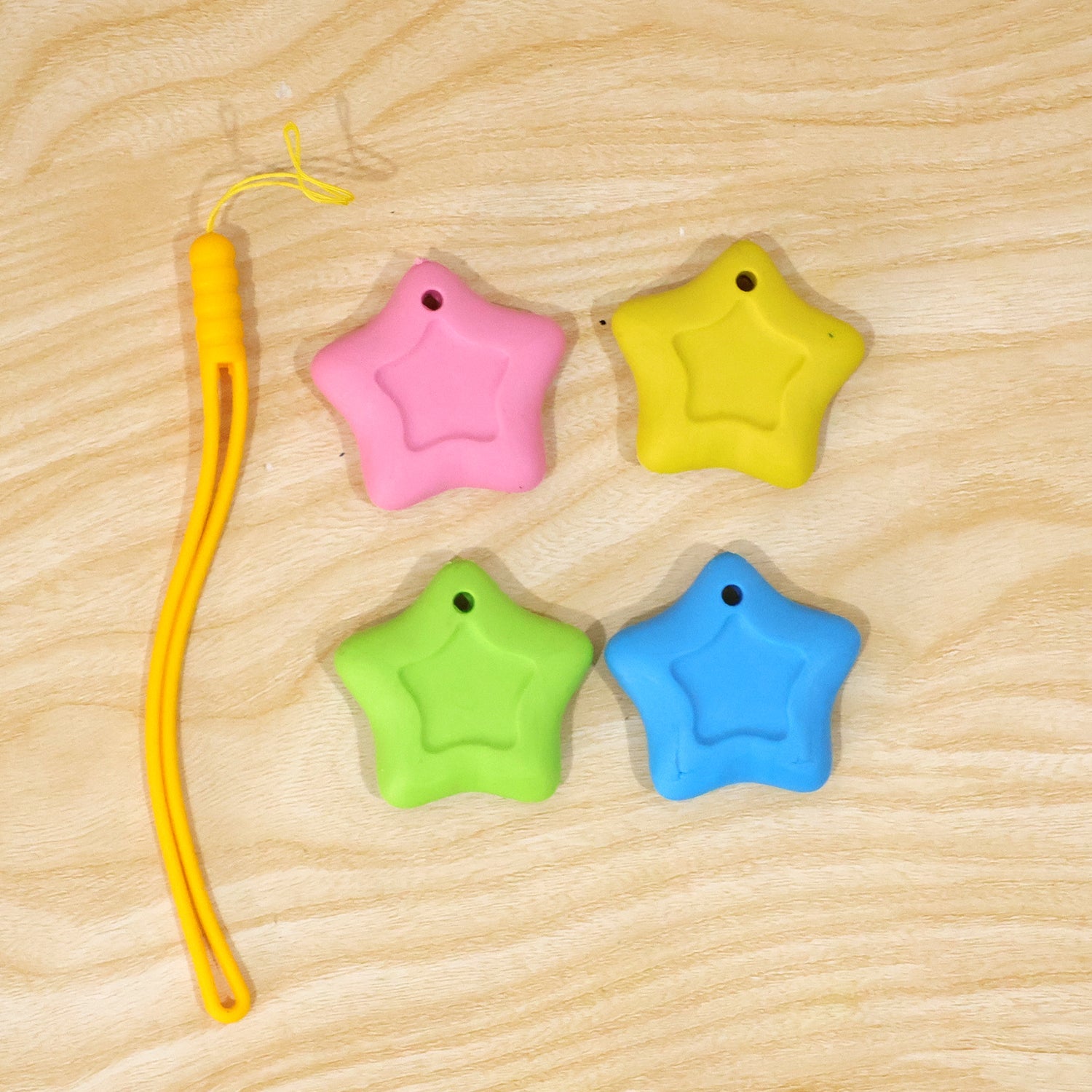 Four star-shaped erasers in pink, yellow, green, and blue are arranged on a light wooden surface next to a yellow lanyard.