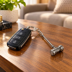 Silver-plated hammer keychain attached to a black car remote, resting on a polished wooden table. A green plant and a sofa