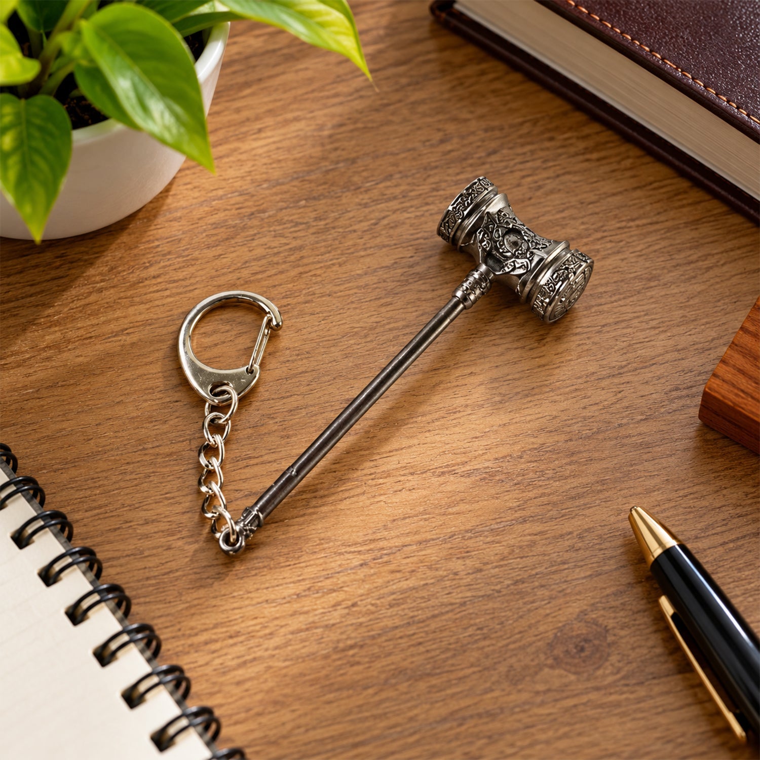 Silver-plated hammer keychain on a wooden desk with a notebook, pen, and a small potted plant.