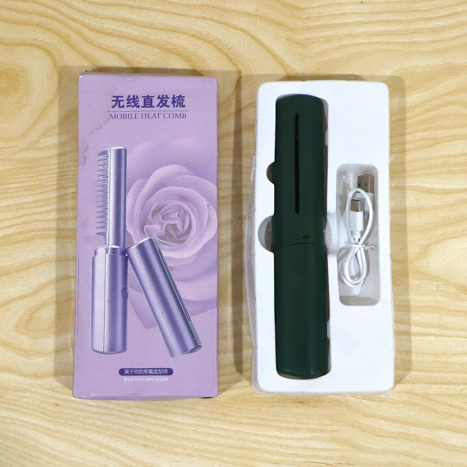 The dark green mobile heat comb and its charging cable are displayed in a white plastic tray next to its purple product box.