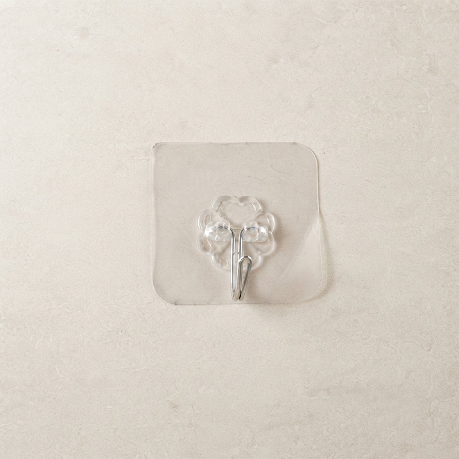 A transparent self-adhesive hook with a silver metal hook centered on a light beige tiled wall.