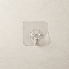 A transparent self-adhesive hook with a silver metal hook centered on a light beige tiled wall.