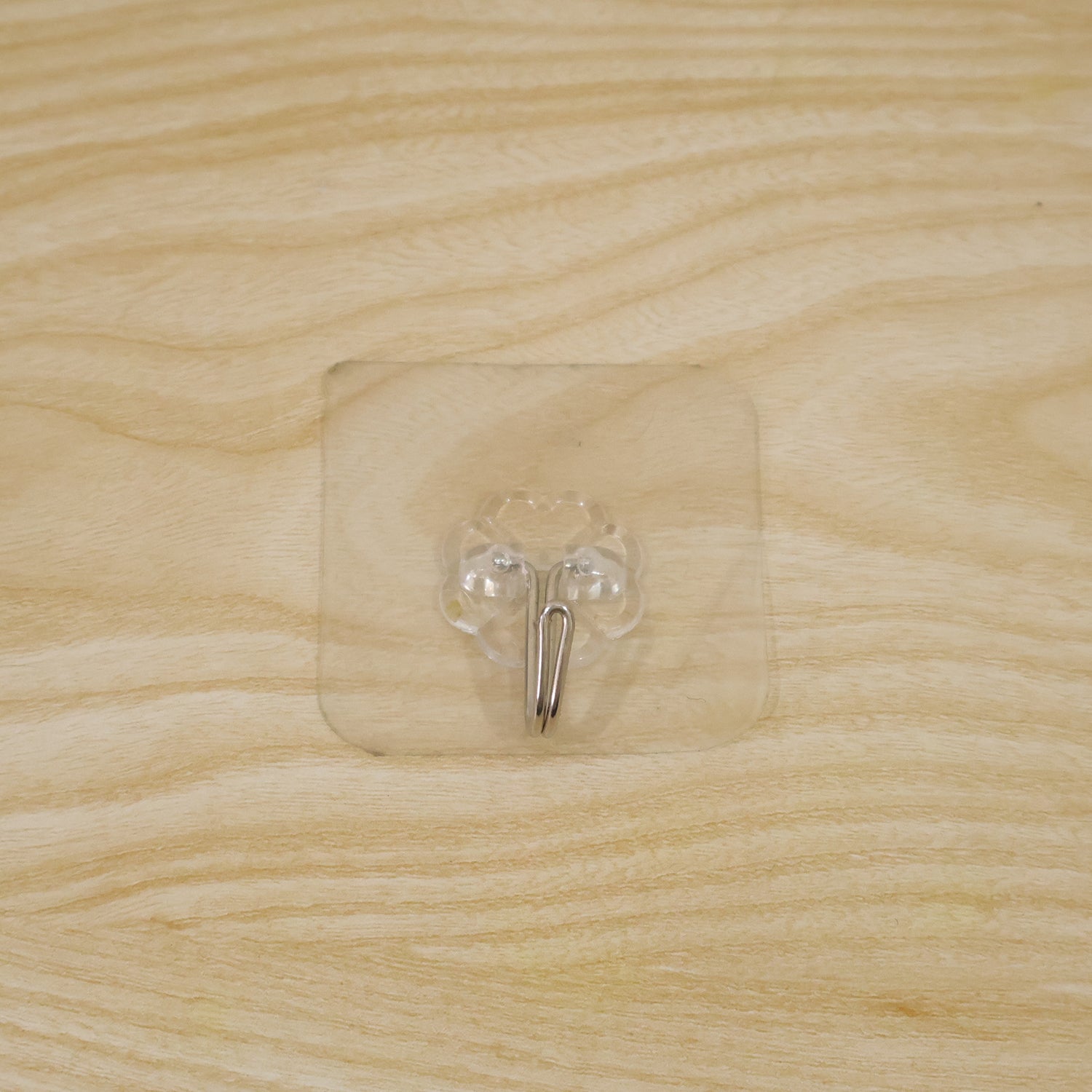 A transparent self-adhesive hook with a silver metal hook centered on a light wooden surface.