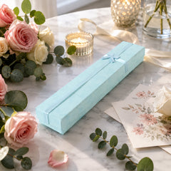 A closed light blue rectangular gift box with a light blue ribbon and bow, on a marble surface with pink roses and candles.
