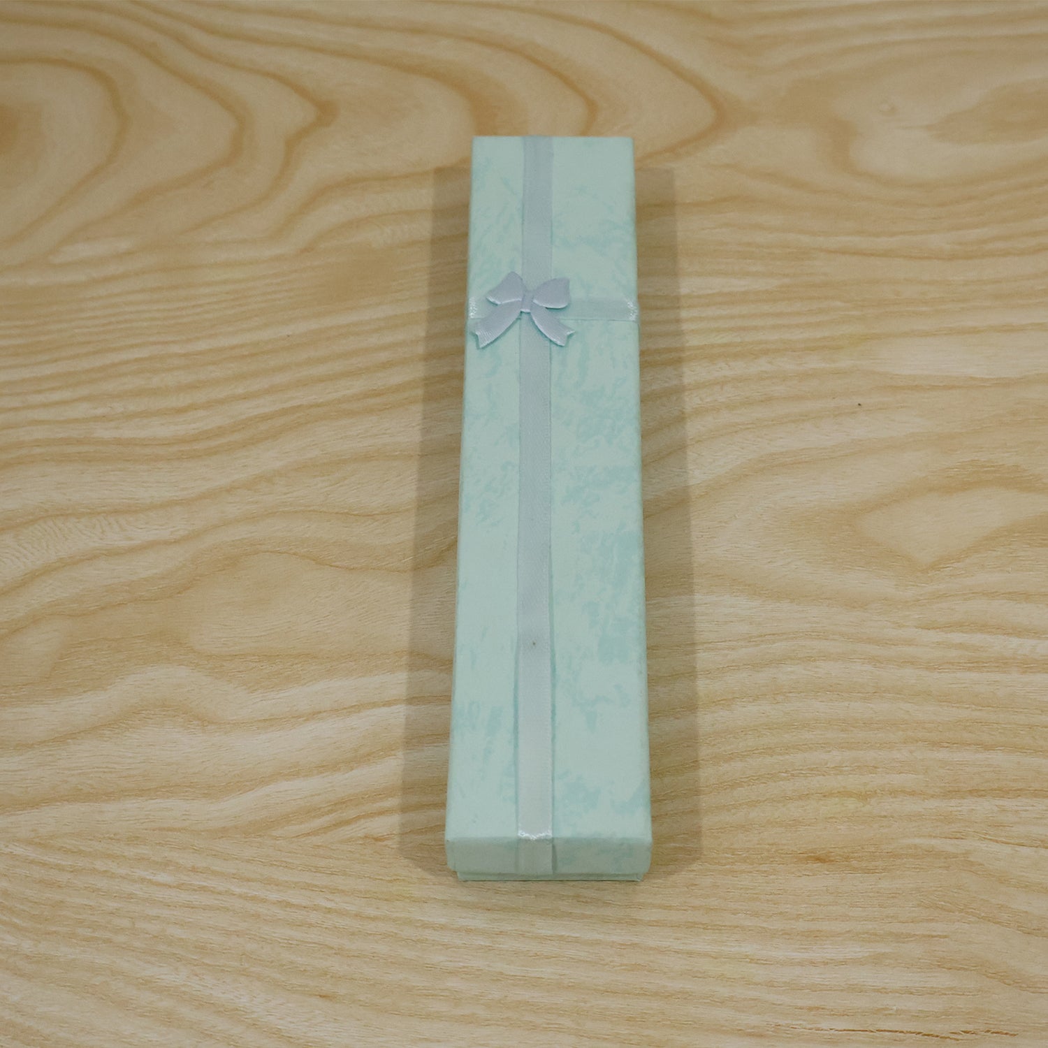 A closed light blue rectangular gift box with a light blue ribbon and bow, on a light wooden surface.