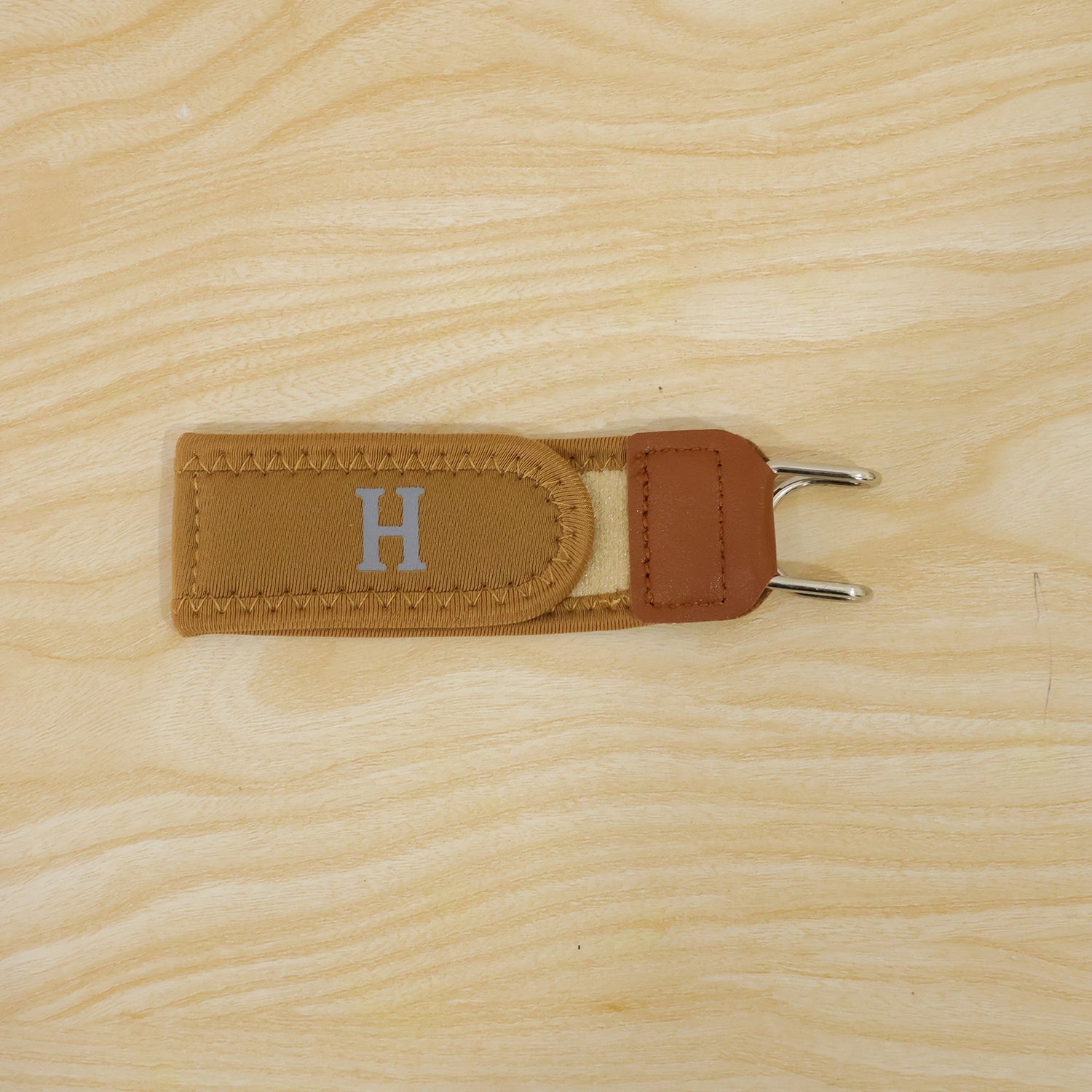 A single tan elastic belt hook with brown faux leather and silver-colored metal hooks, centered on a light wooden surface.