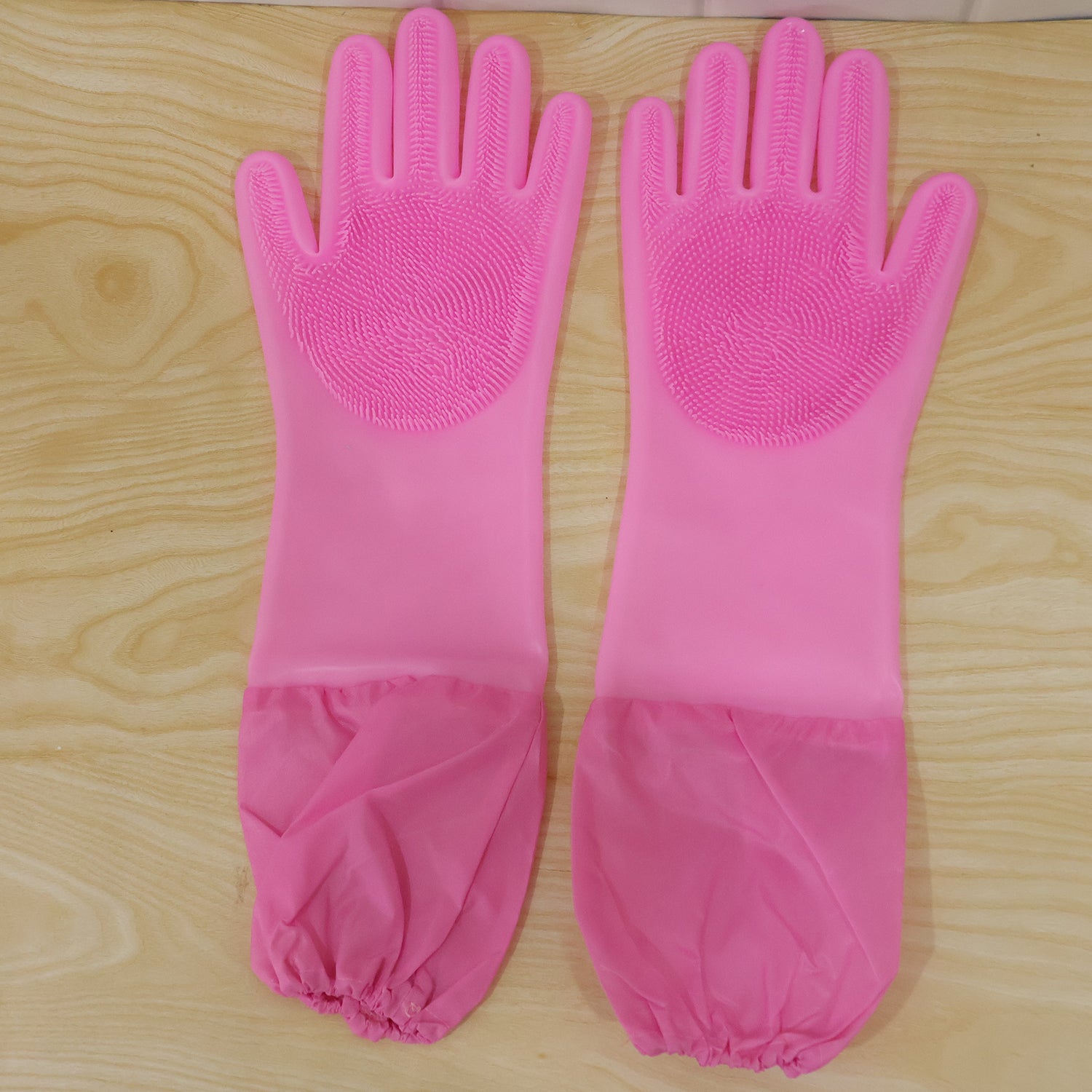 A pair of pink silicone scrubbing gloves with long, gathered cuffs laid flat on a light wooden surface.