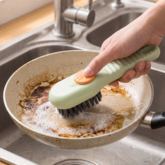 A hand uses a green and orange liquid-dispensing brush to clean a dirty frying pan in a stainless steel sink.
