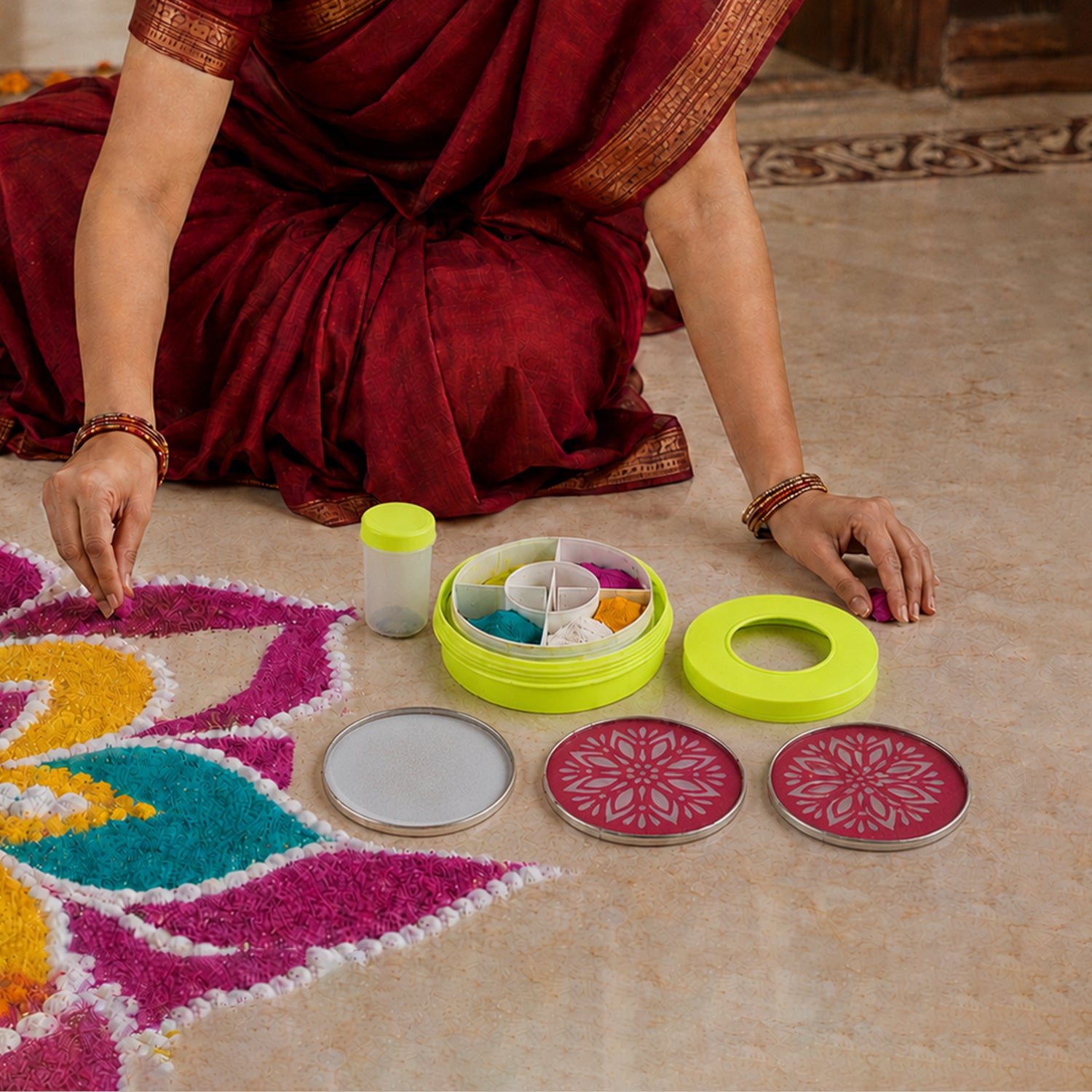 A person sits on the floor, surrounded by a partially completed colorful rangoli, stencils, and a powder dispenser.