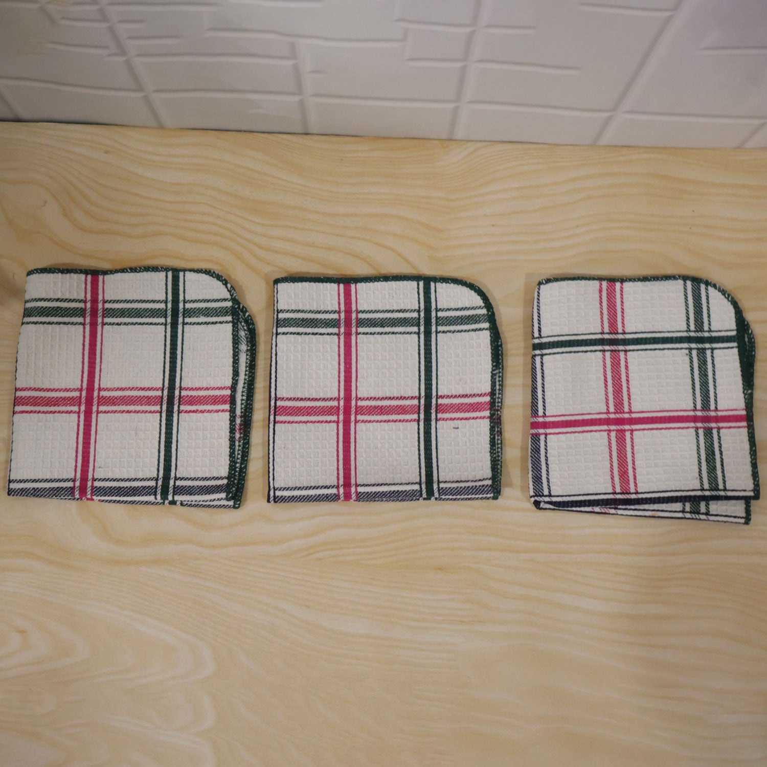 Three plaid dishcloths, each folded into a square, are laid side by side on a light wooden surface.