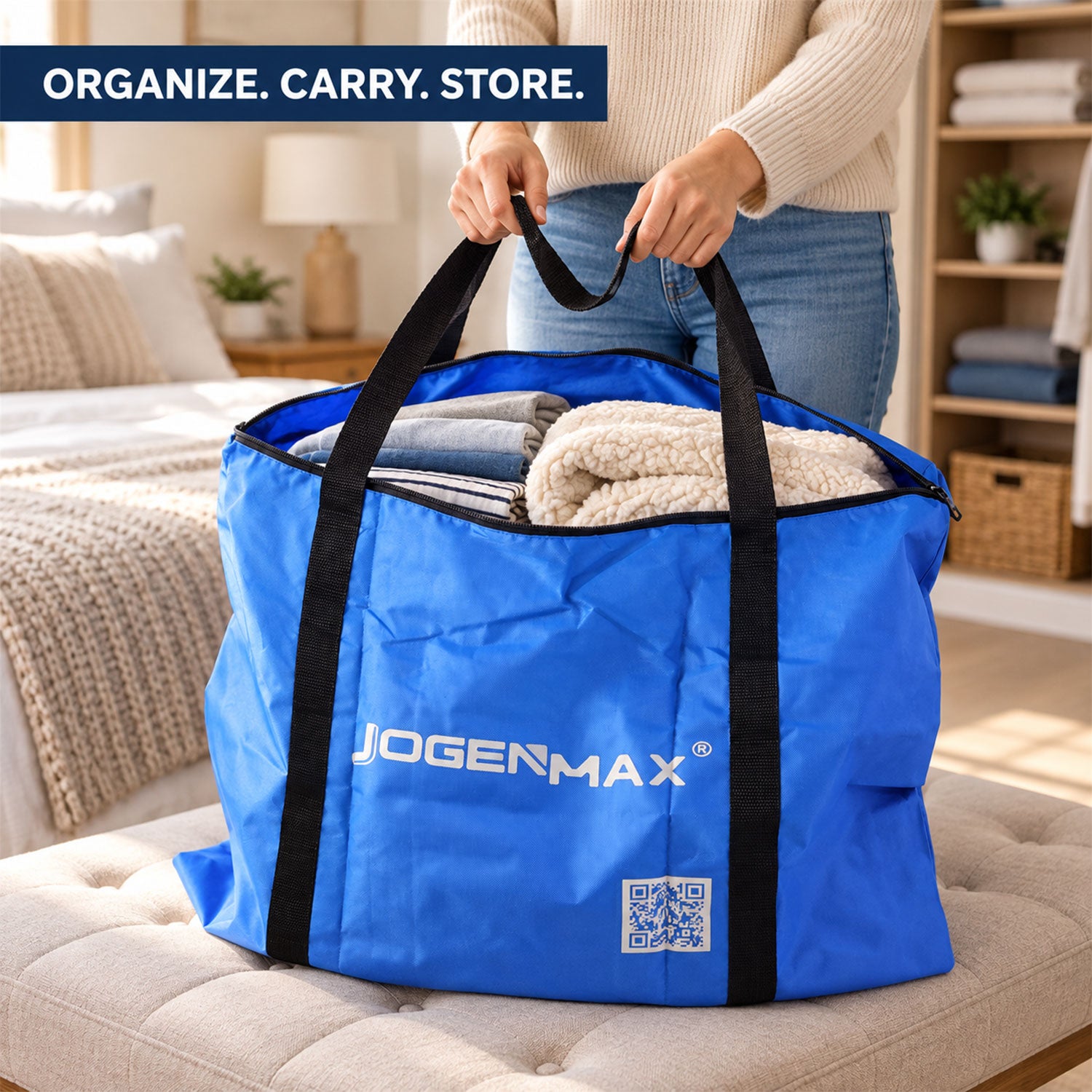 A person holds a blue JOGENMAX storage bag filled with clothes and blankets by its black handles on a cushioned bench.