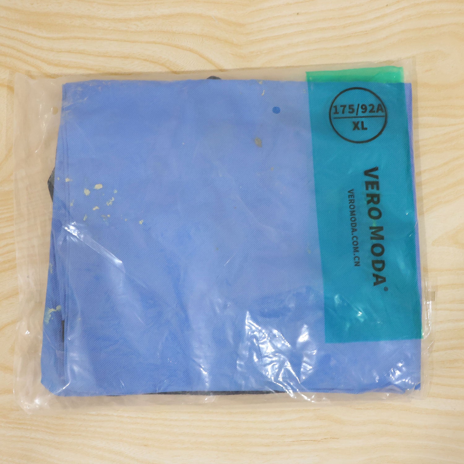 A blue folded item, possibly the bag, is sealed inside a clear plastic package with a green and white label.