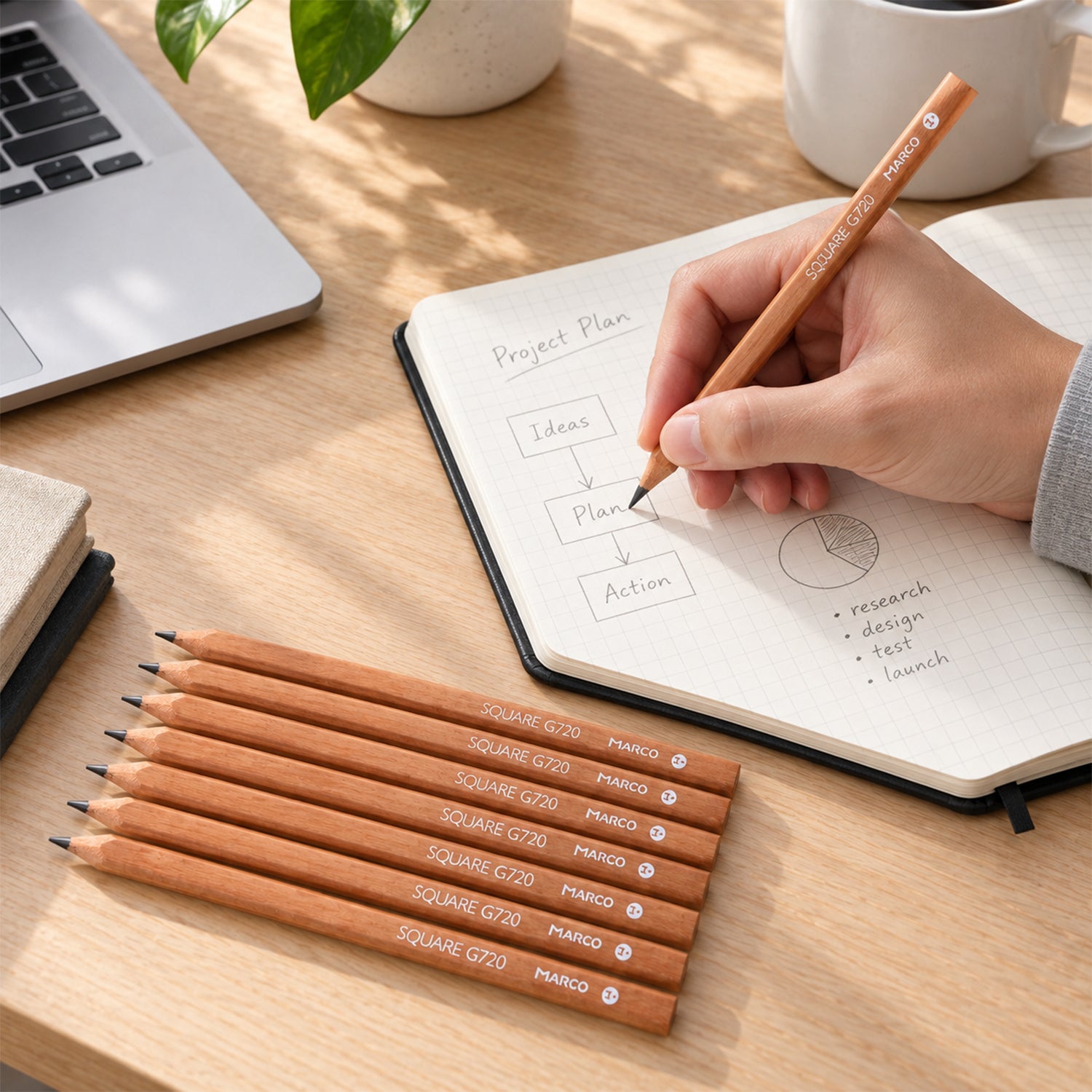 A hand writes in a notebook with a light brown square pencil, surrounded by seven similar pencils and a laptop.