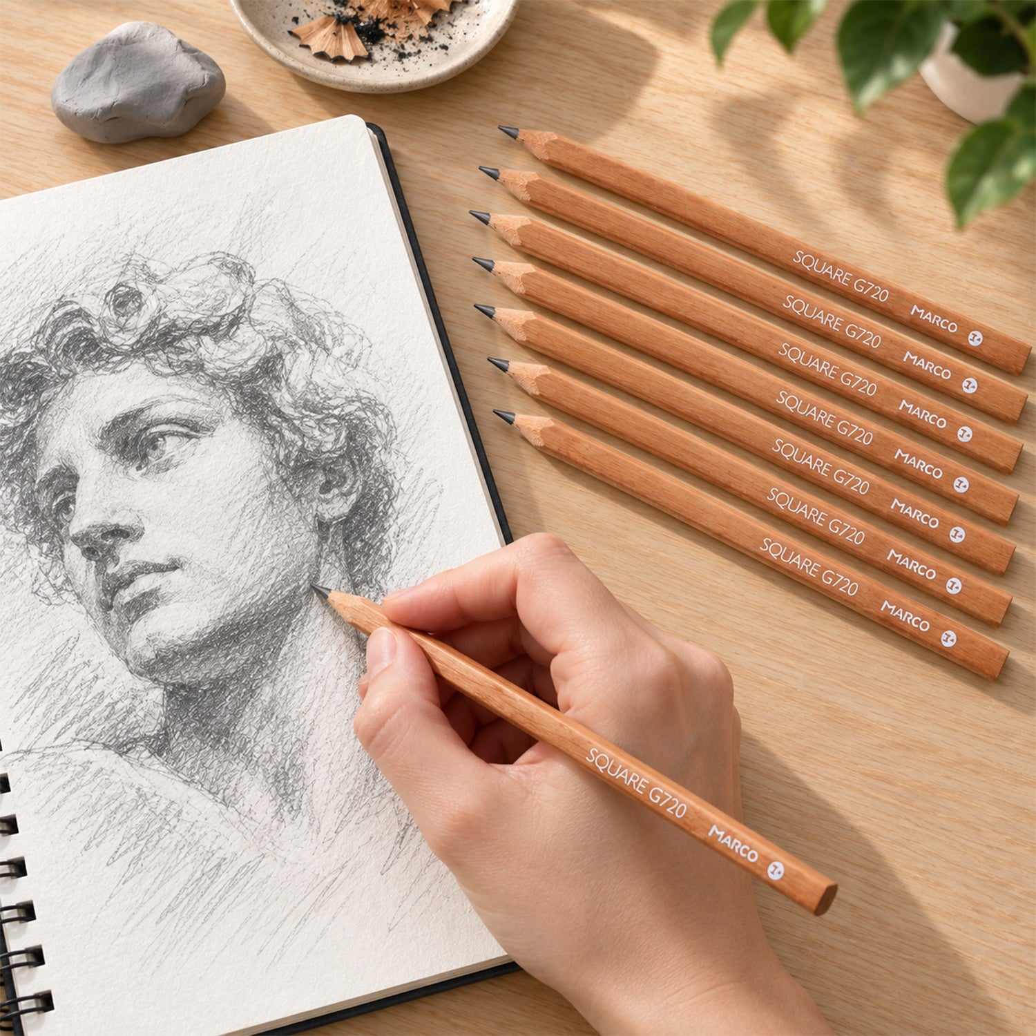 A hand sketches a classical bust in a spiral-bound notebook using a light brown pencil, with seven other pencils beside it.