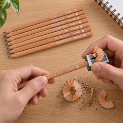 Two hands sharpen a light brown pencil with a metal sharpener, creating wood shavings on a wooden desk next to seven other