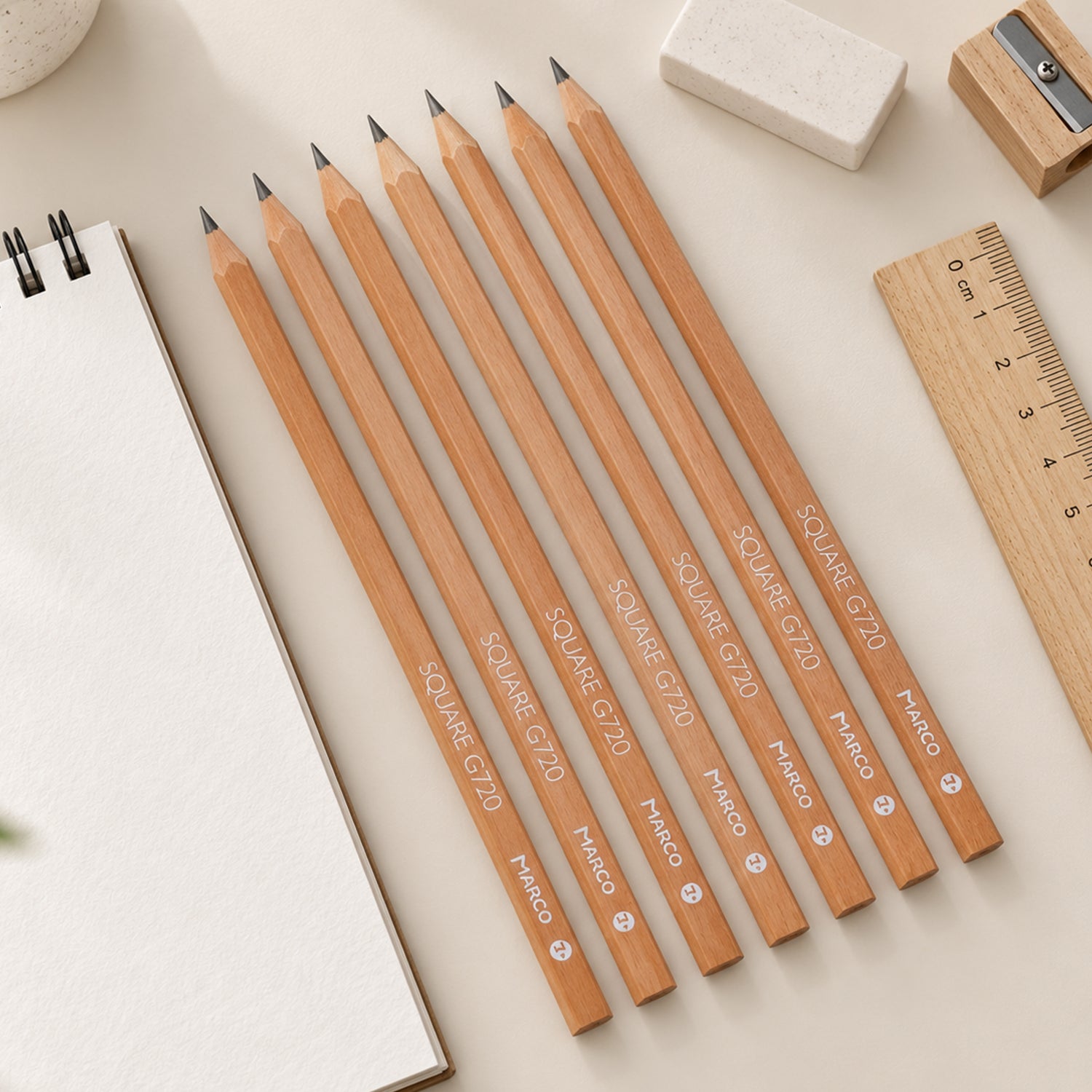 Eight light brown square pencils are fanned out on a light-colored desk beside a spiral notebook, eraser, and ruler.