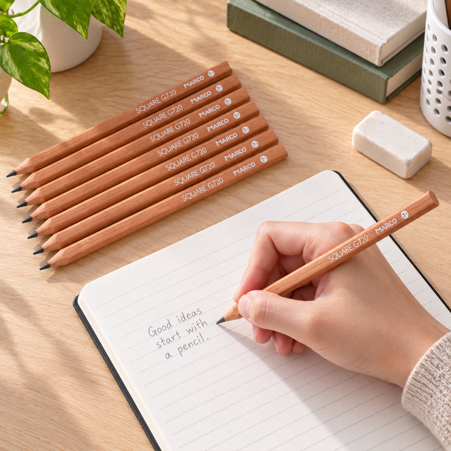 A hand writes in a notebook with a light brown pencil, surrounded by seven identical pencils, an eraser, and a plant.