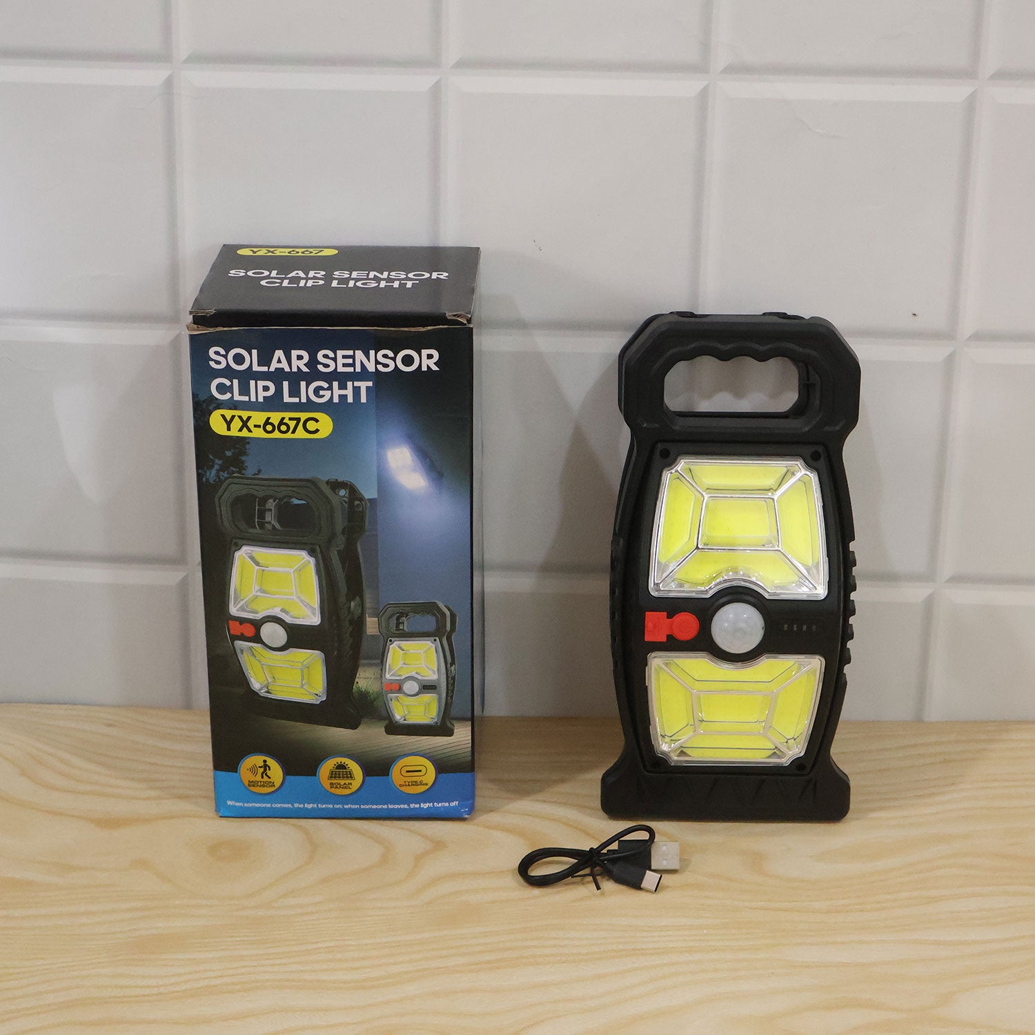 A black solar sensor clip light, its retail box, and a USB-C cable are displayed on a wooden surface against a tiled wall.