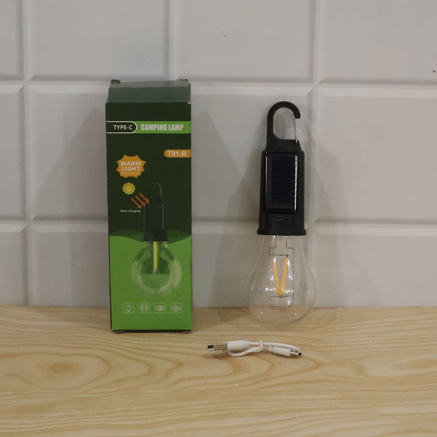 The solar bulb light, its green retail box, and a white USB-C charging cable rest on a light wood surface against a tiled