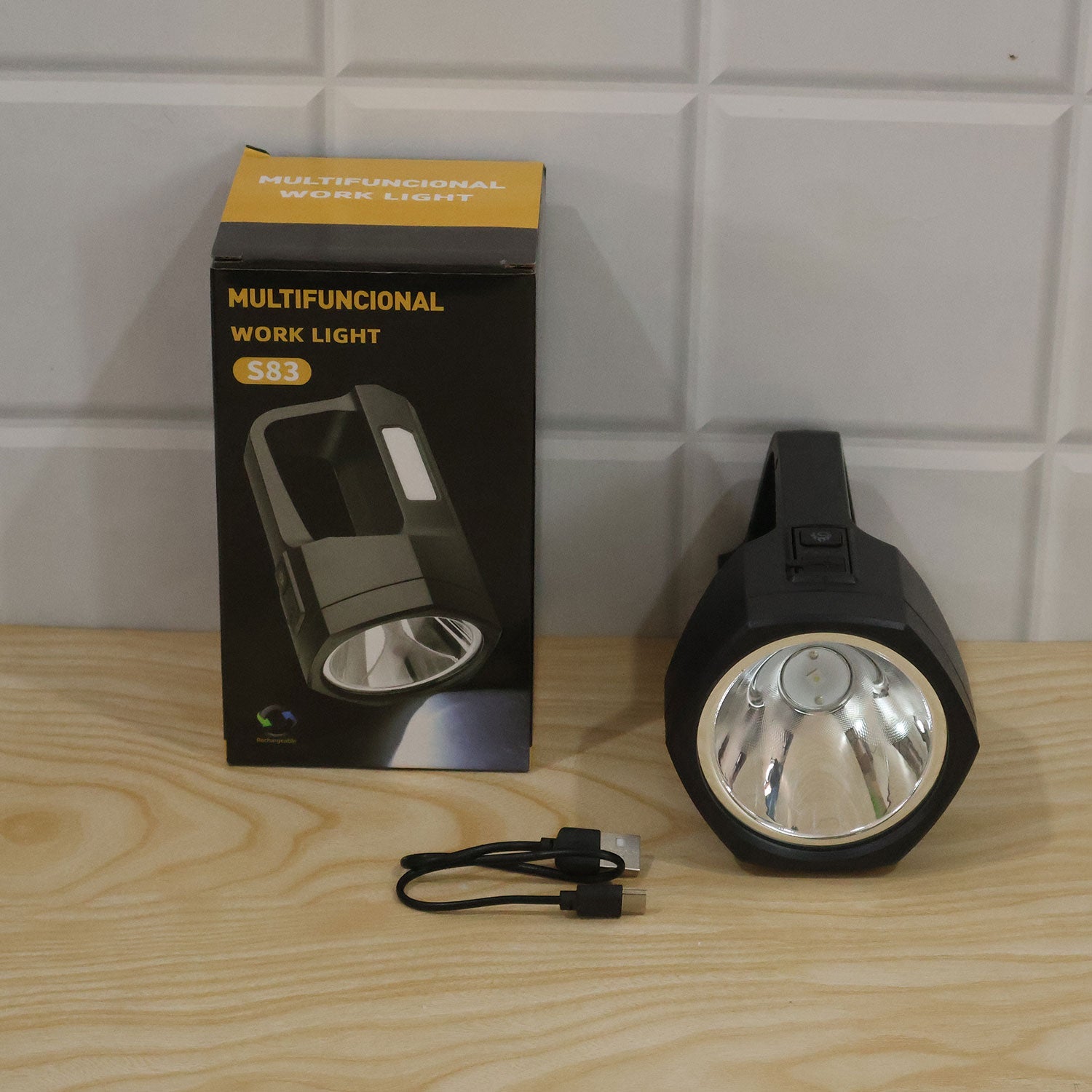 A black torch, its packaging box, and a USB-C cable resting on a light wooden surface against a white tiled wall.