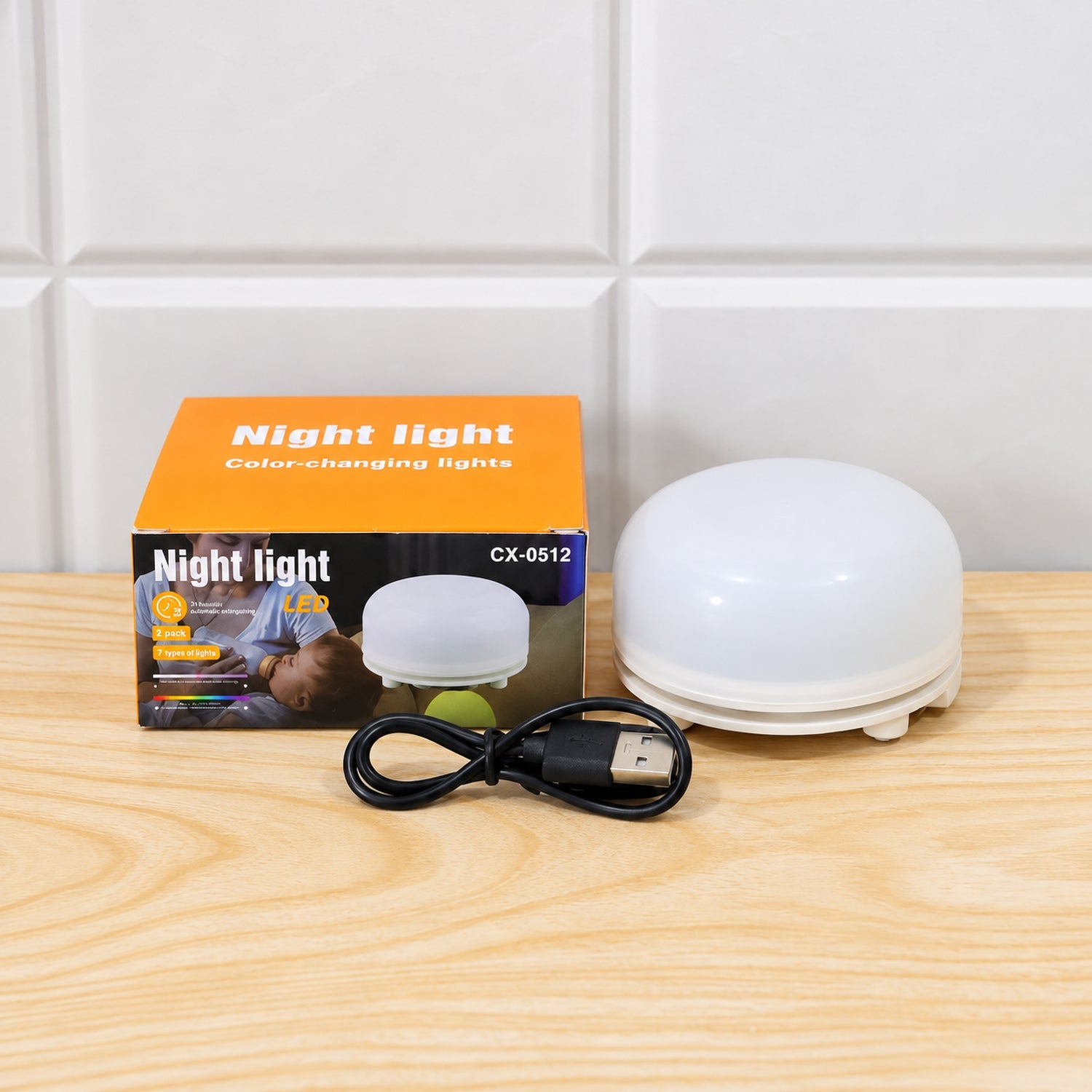 A white dome-shaped night light, its orange box, and a black USB cable are arranged on a light wooden surface.
