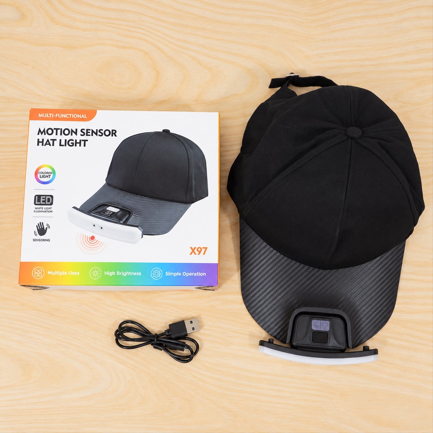 A black baseball cap with an LED light, its product box, and a coiled USB charging cable laid out on a light wooden surface.