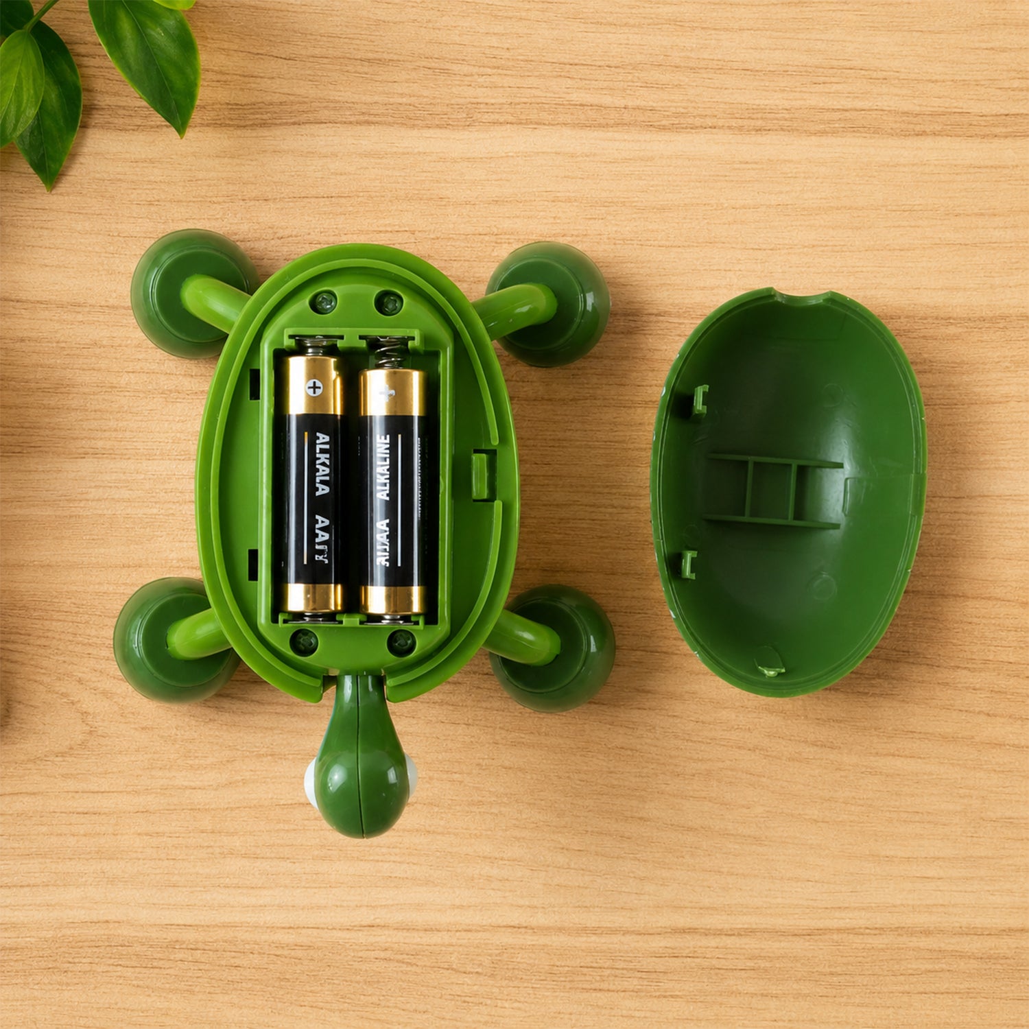 The green turtle massager is open, revealing two AA batteries inside, resting on a wooden surface next to its shell.